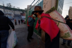Por el COVID19 mucha gente dejo de ir al mercado San Roque y prefiere de comprar los productos en los supermercados. Ironía: muchas de las frutas, verduras y granos que se compra allí, provienen del mercado en el Centro de Quito. – Una mujer saliendo de una bodega con hierbas en la espalda, mascarilla en la cara y guantes en las manos.
